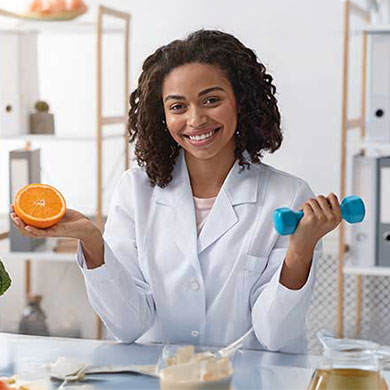 a sports nutritionist smiling at the camera holds an orange in one hand and a weight in the other